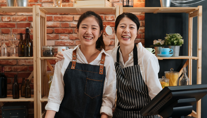 Two woman in aprons smile to the camera from behind a retail counter.