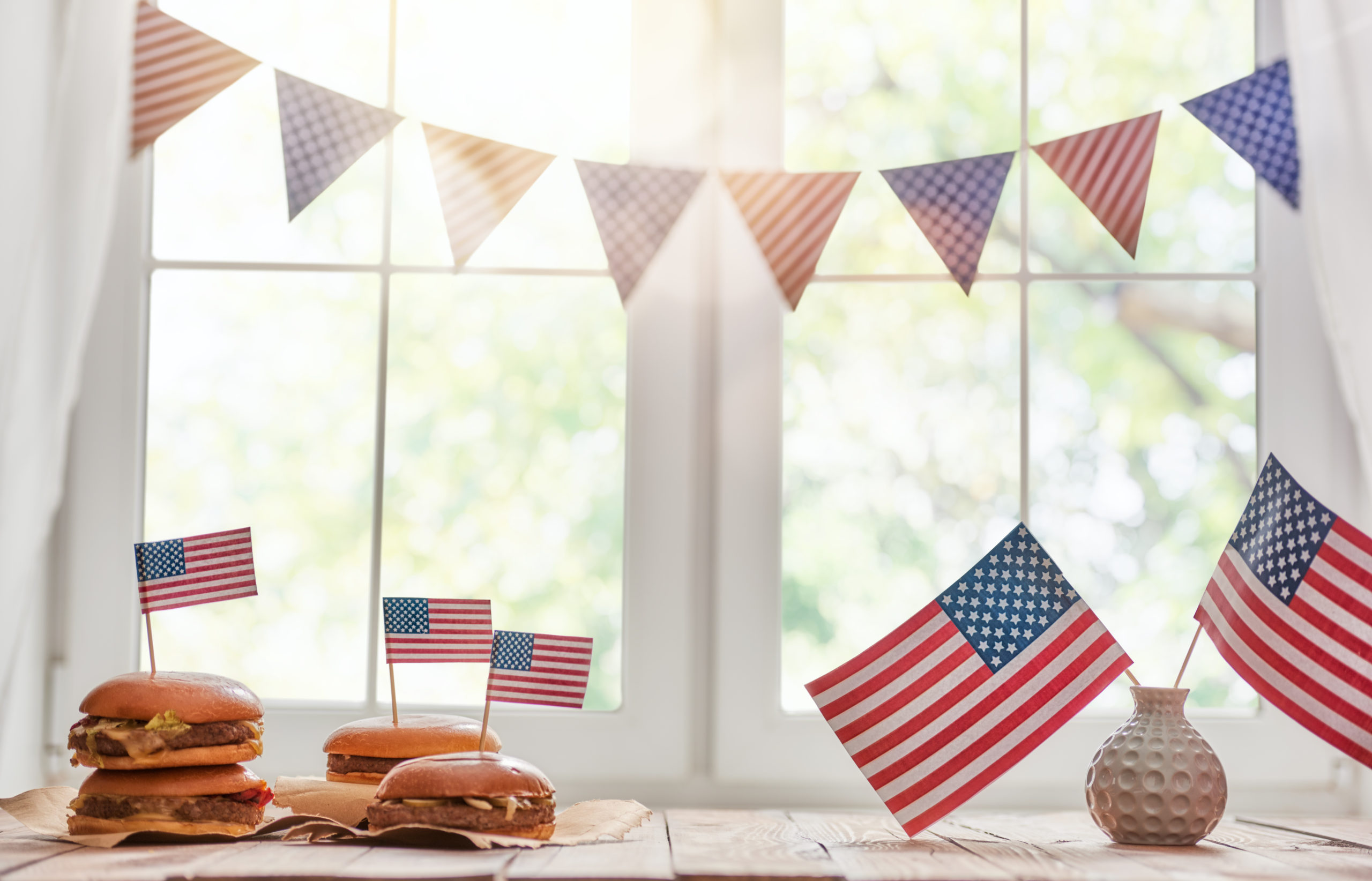 Various foods sit on a plate in a window with American flags sticking out. There is vase to the right also holding American flags and bunting above in read and blue.