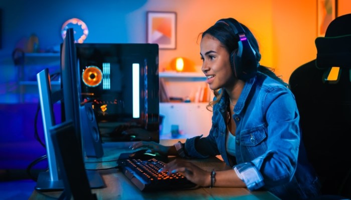 Gamer girl in headphones playing a video game online on her computer in a room with colorful lights.