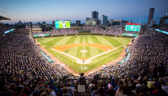 A packed baseball stadium during an evening game, with players on the field and large screens displaying content. The skyline of the city is visible in the background.