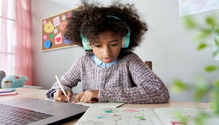A young girl works at her desk in front of her computer with headphones on.
