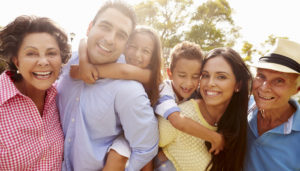 A couple with their kids hugging them on their backs smiles at the camera, a grandparent smiling on either side as well.