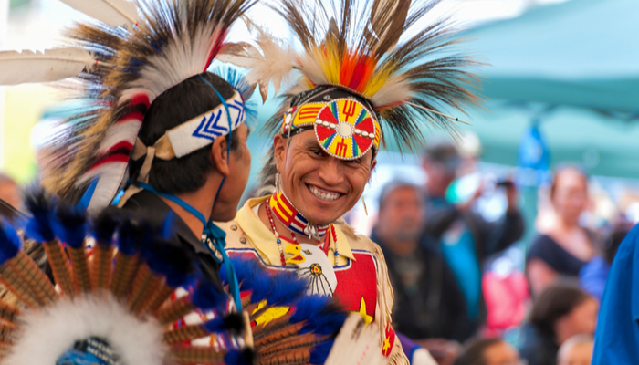 To men smile at each other while dressed in traditional Native American attire.