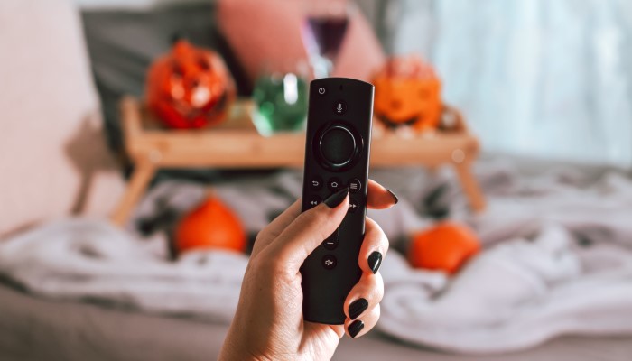 A close-up of a woman's hand holding up a television remote. Halloween decor and party favors can be seen in the background.