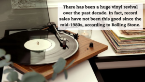 A turntable with vinyl record sitting on desk next to a stack of records.