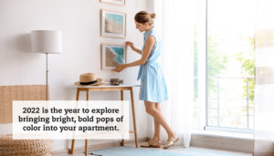 A woman hangs a painting on her wall in a bright, sunny room. A quote reads: "2022 is the year to explore bringing bright, bold pops of color into your apartment."