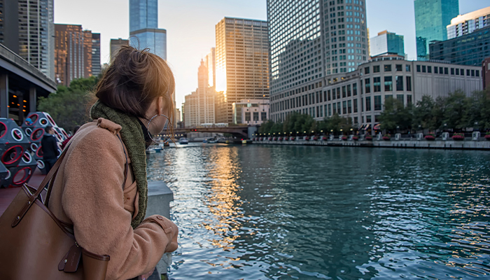 A woman leans on a railing overlooking the Chicago River at sunset. There are buildings along the riverbank on the far side.