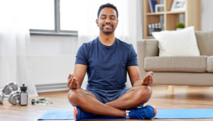 A man sits on a yoga mat in his living room sitting cross-legged, meditating.