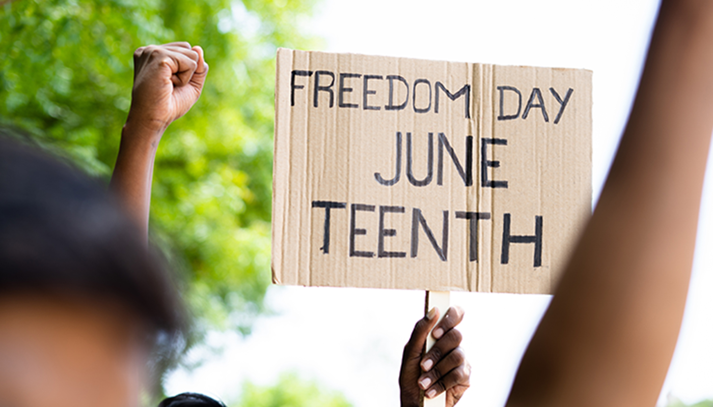A close-up of hands in the air at and event outdoors. One hand is holding a sign that says "Freedom Day June Teenth"