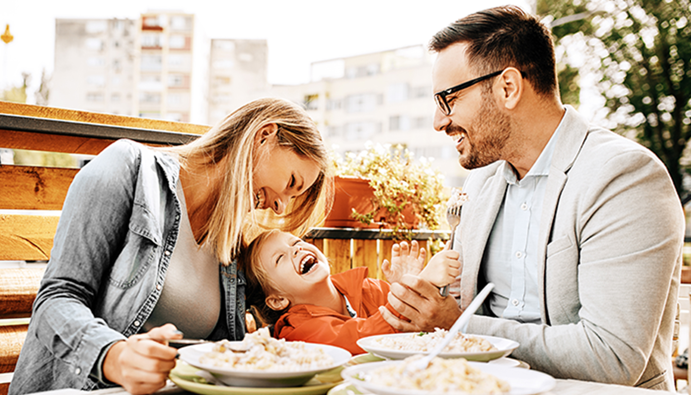 A family having a meal at an outdoor café. The father smiles at his son who is laughing and leaning against his mother.