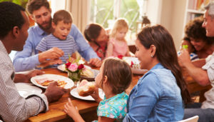 A family sits around a dining table sharing a meal. On the right a couple hugs from the chairs and three other parents have small children in their laps.