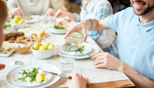 A close-up of a man pouring water into someone's glass at a dining table that salads and bread on it.