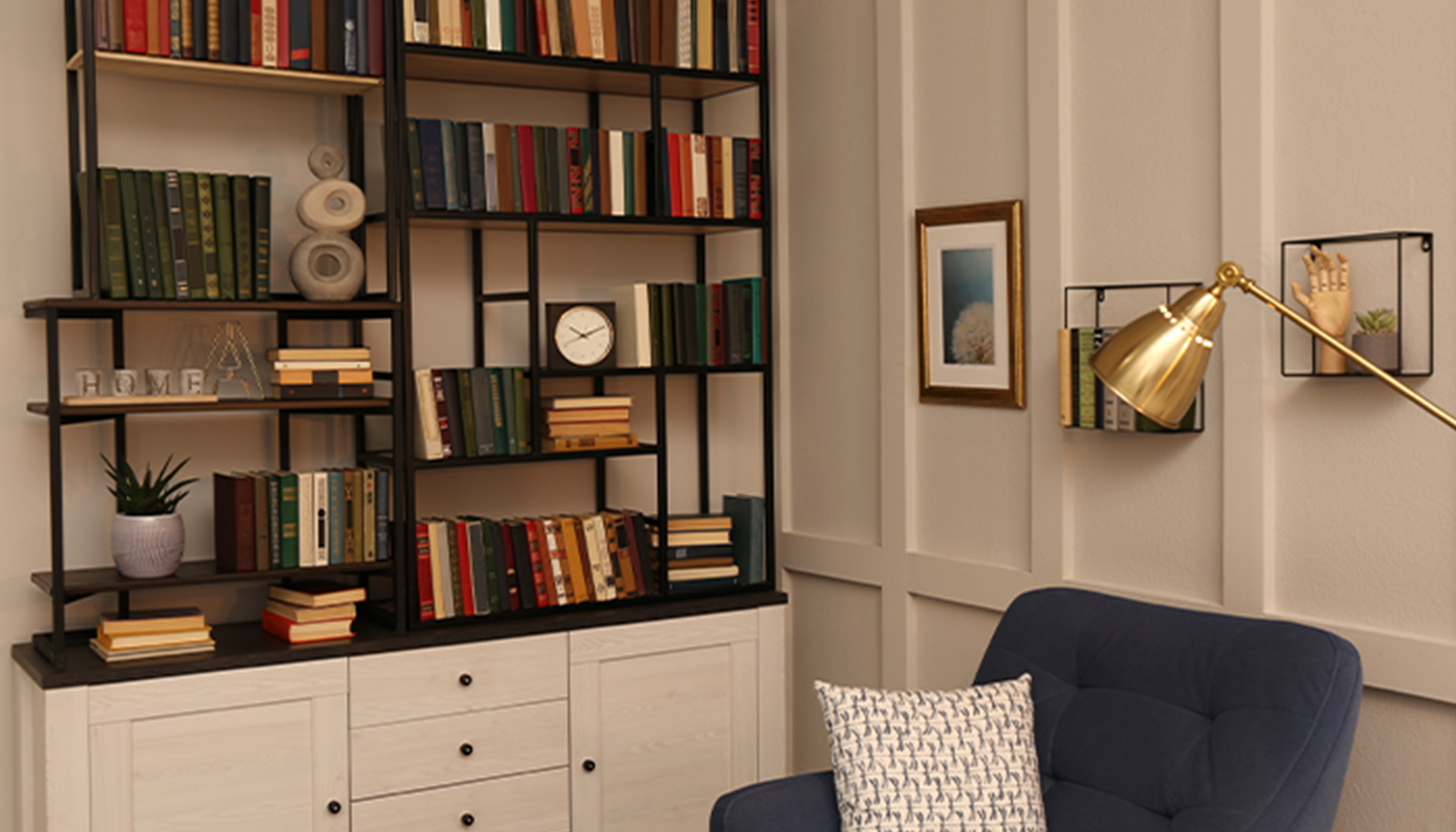 Looking at the corner of a living room. On the right is a white panel wall with pictures hanging over a navy chair and brass lamp. On the left is a nearly full bookcase atop a white dresser.