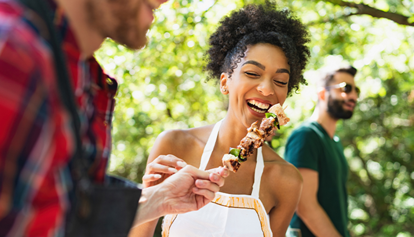 A woman laughs as a man hands her a chicken kabob right off the grill. There is another man behind her wearing sunglasses. They are all outdoors under several trees.