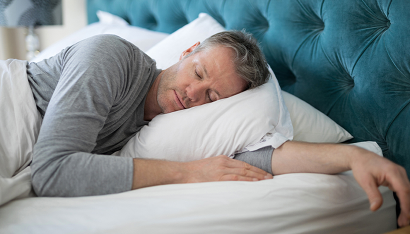 A man sleeps on his side facing the camera with one are under the pillow. He's in a grey shirt under white covers. The headboard is blue.