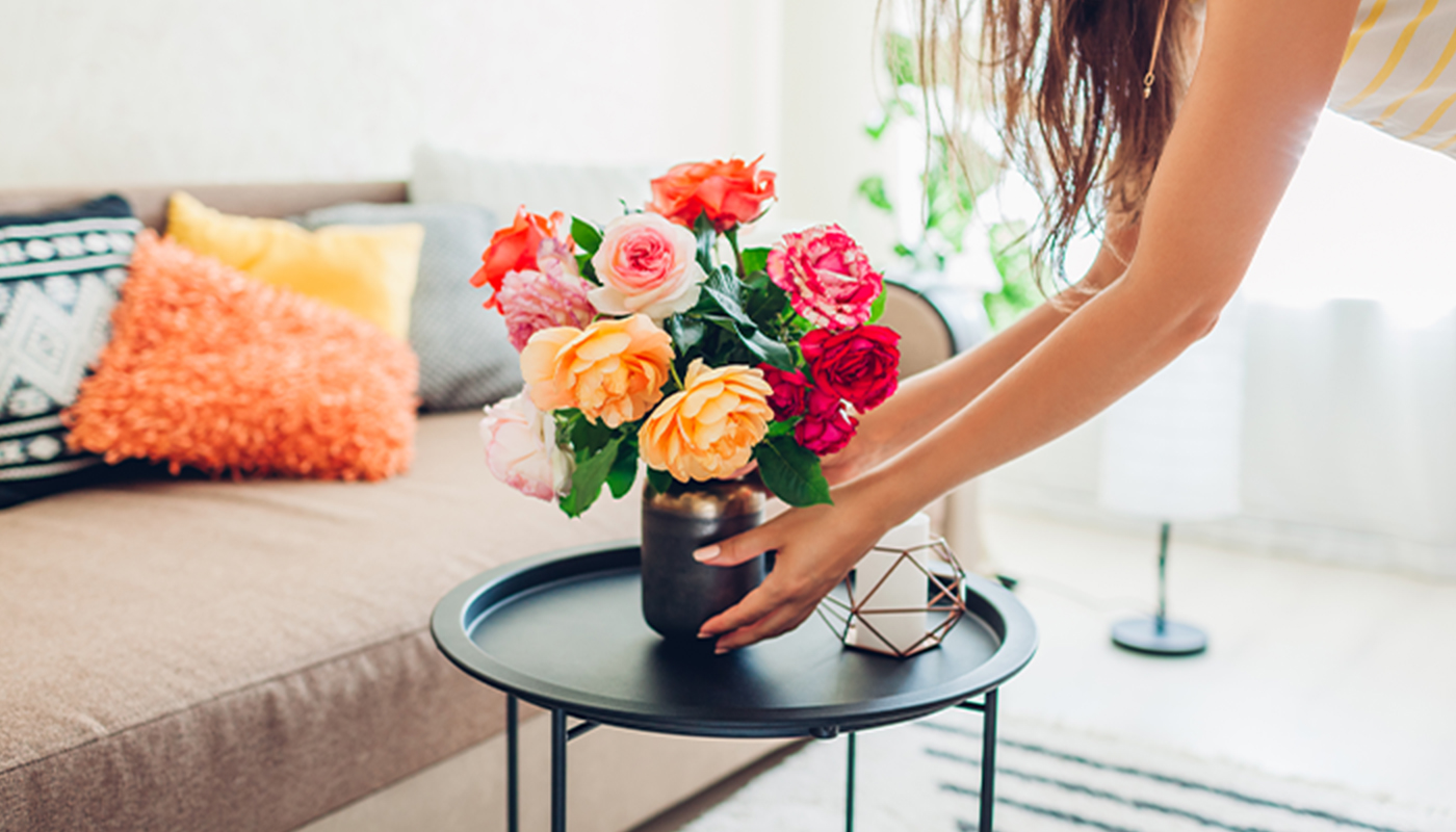 A woman arranges a bouquet of red, pink and orange roses in a vase set atop a small, round table in her living room.