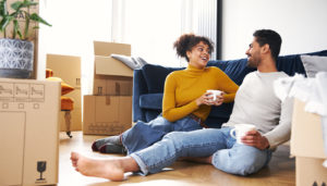 A couple sits on their floor, leaning on a couch taking a coffee break from unpacking boxes which are all around them.