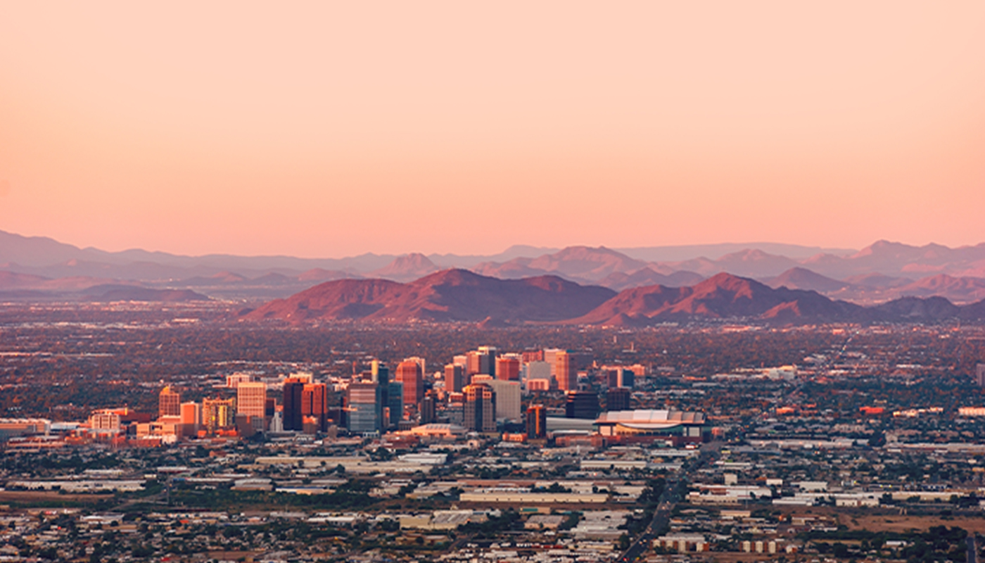 Looking at downtown Phoenix from a very far distance. The downtown area is seen clustered together in the middle with smaller buildings surrounding it. Mountains are seen in the distance at sunset.