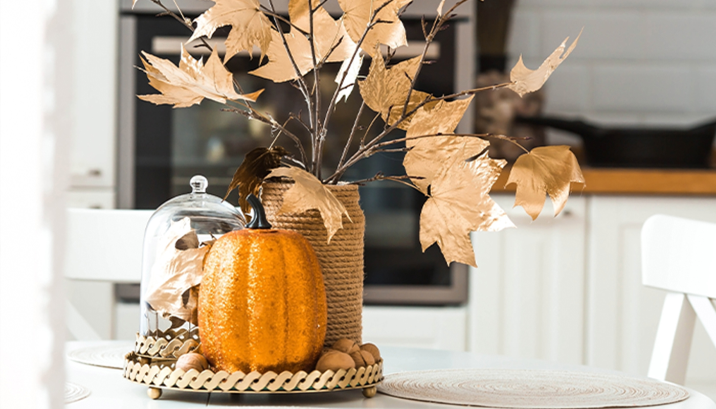 A close-up of fall décor on a table, which include a pumpkin and a vase holding orange leaves.