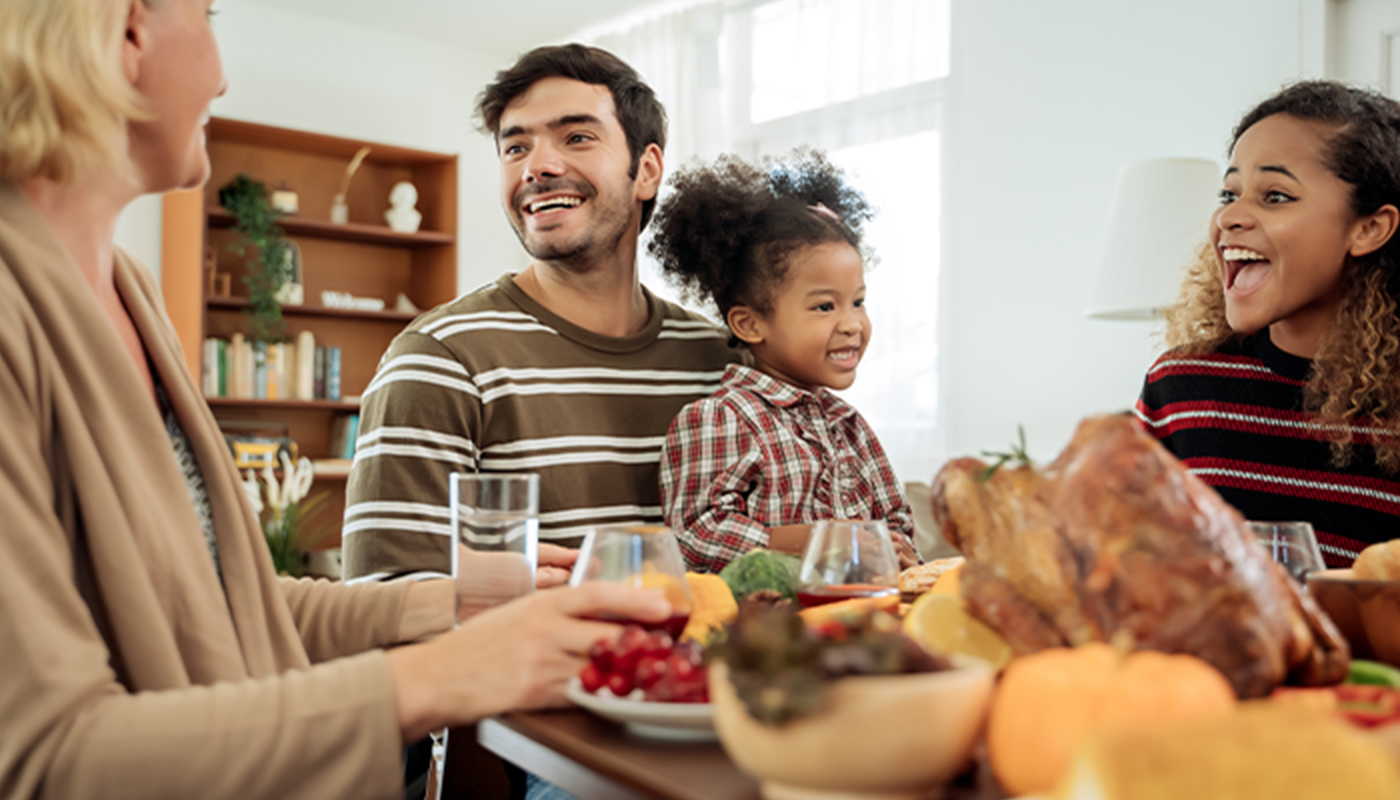 A family sits around a Thanksgiving table. A man has a child in his lap. People are talking and smiling. There is a lot of food on the table.