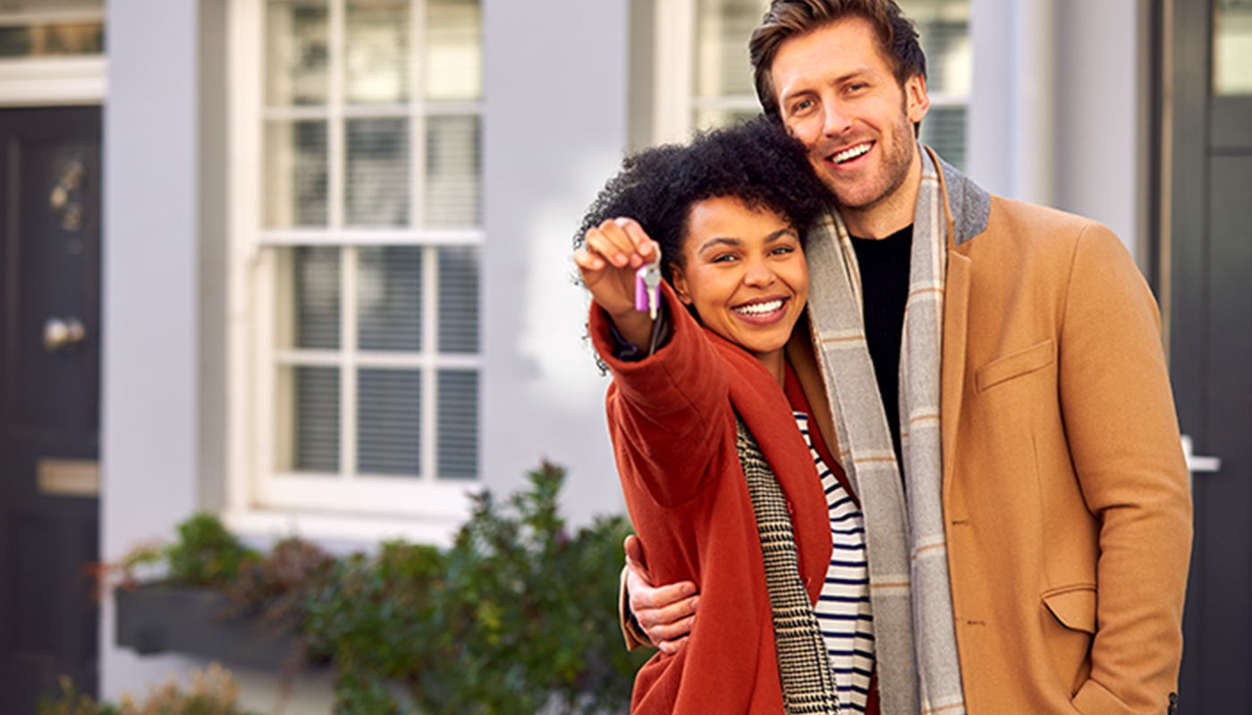 A couple smiles holding up the keys to their new house. They are standing outside, in front of the house, wearing winter jackets.