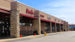 Looking from street level across front of the storefront of Pickwick Place Shopping center. There is a covered sidewalk on the left and the parking lot is on the right.