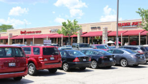 Looking from street level across the parking at the storefront of Pickwick Place Shopping center. There is a row cars in the foreground. The storefront is behind in the background.