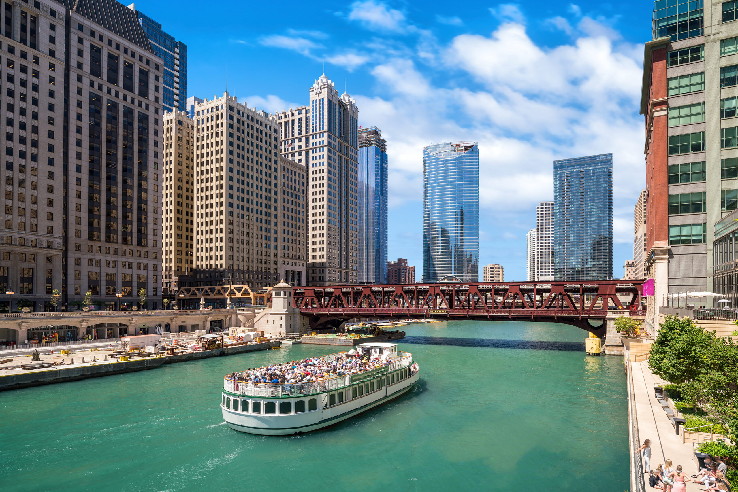 A double-decker boat, open on the top, drives people down the Chicago River through downtown on a sunny day. Building rise on both sides of the river.