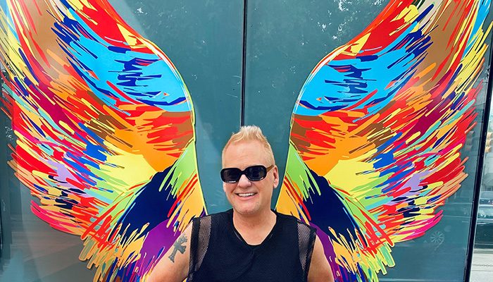 A portrait of Richard Melton standing before bright and colorful wings painted on a wall.