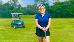 Brenda standing on a golf course holding a putter on a sunny day. There is a golf cart parked behind her in the background.