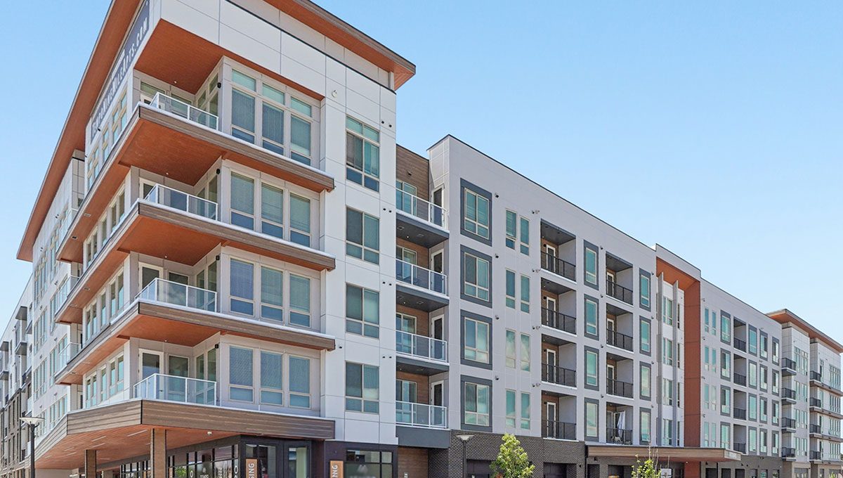 Modern mid-rise apartment building with large windows, private balconies, and mixed exterior materials including glass, metal, and wood accents under a clear blue sky.