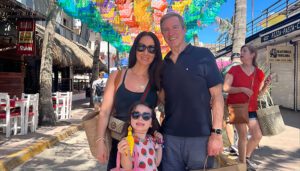 A family of three, with a man, a woman, and a young girl holding a lollipop, posing together on a sunny street with colorful decorations overhead.