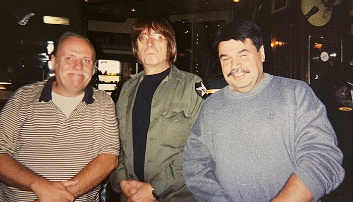 Three men standing together in a bar, smiling; one in a striped shirt, one in a green jacket, and one in a gray sweater.