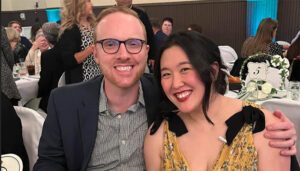 A man and woman are seated together at a banquet event, smiling at the camera. The man wears glasses and a dark jacket, while the woman is in a yellow patterned dress. Other attendees are visible in the background.