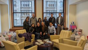 A group of ten people standing and seated in a room with large windows. Several bags and wrapped gifts are on the floor around them.