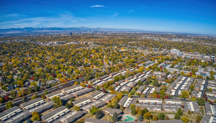 Aerial view of a sprawling cityscape with suburban neighborhoods, tree-lined streets, and distant city buildings against a clear blue sky.