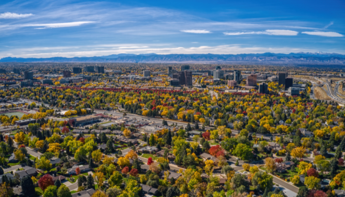Aerial view of a city with a mix of buildings and trees in various fall colors, set against a backdrop of mountains under a clear blue sky.