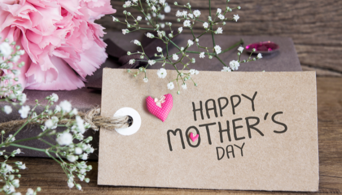 A mother's day card with "happy mother's day" written on it, adorned with a pink heart, alongside pink flowers and a small white flower bouquet on a wooden surface.