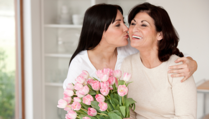 A younger woman kissing an older woman on the cheek, both smiling, holding a bouquet of pink tulips indoors.