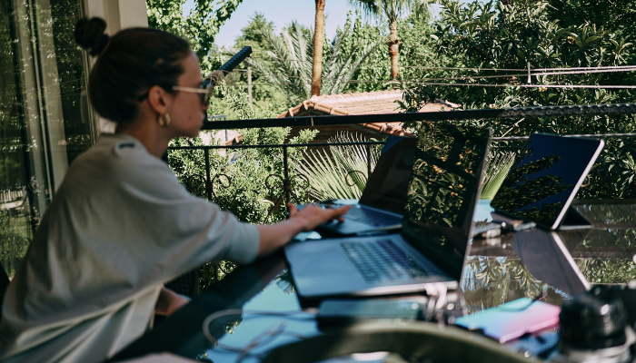 A person works on a laptop and other devices on an outdoor patio with a view of tropical foliage.