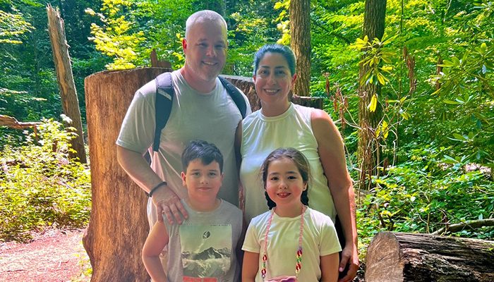 A family of four stands together in a forest, smiling at the camera. Two adults are behind two children, with greenery and trees visible in the background.