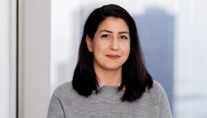 A woman with shoulder-length dark hair, wearing a light gray sweater over a pink collared shirt, stands in front of a window with an urban cityscape in the background.