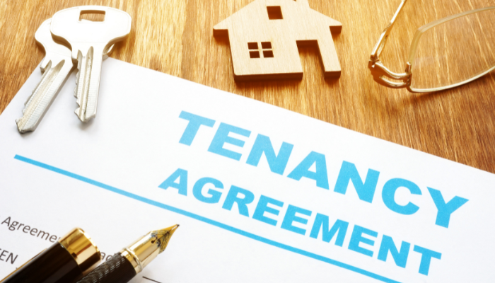 A tenancy agreement document on a wooden surface, with a pen, keys, a house-shaped keychain, and a pair of glasses.