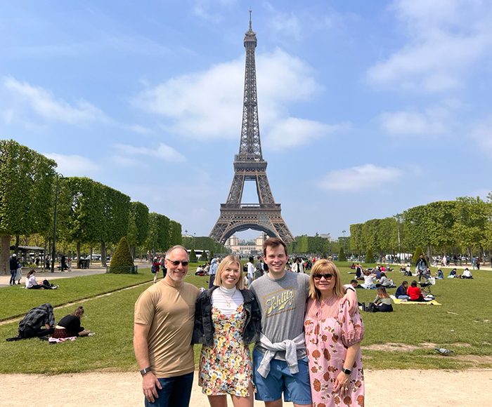 Four people stand together smiling on a grassy area in front of the Eiffel Tower, with scattered people sitting on the grass under a blue sky.