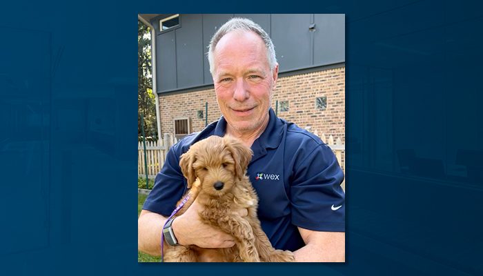 Man wearing a navy blue shirt with a logo, standing outdoors and holding a fluffy brown puppy. Brick building and fence are visible in the background.