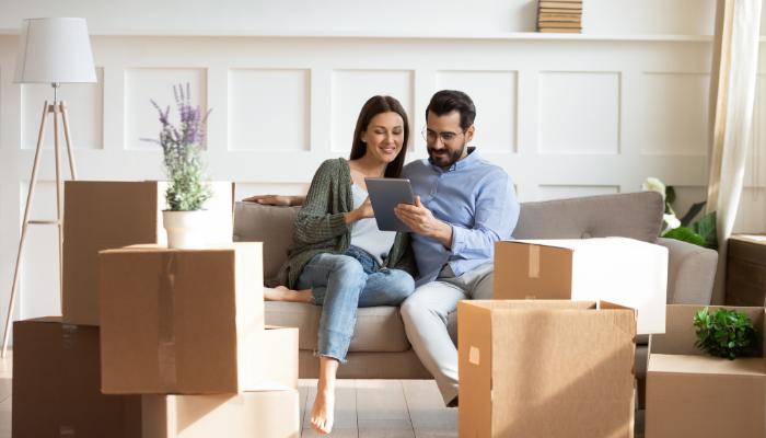 A couple sits on a couch surrounded by moving boxes, looking at a tablet together in a bright, white room with a potted plant and a standing lamp.