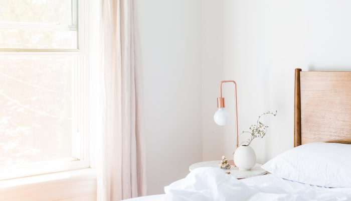 A minimalist bedroom features a wooden headboard, a white bed with white bedding, a bedside table with a vase, and a lamp. Light streams in through a window with sheer curtains.