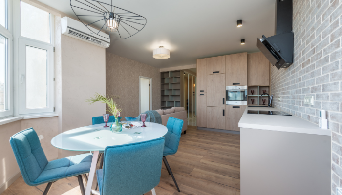 Modern kitchen and dining area with wooden floors, blue chairs around a white table, built-in appliances, gray brick walls, and large windows letting in natural light.