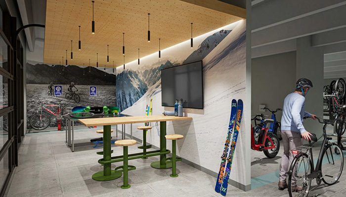 Room with a large mural, wooden table, green stools, bicycles, ski equipment, a wall-mounted TV, and a man wearing a helmet and facing bike racks.