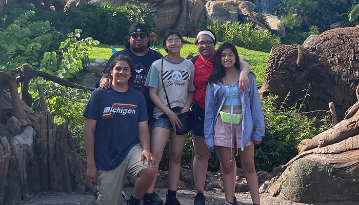 Elizabeth Cañongo stands with her family outdoors in a lush, wooded setting. She is wearing a red shirt and has her arm around her daughter, who is wearing a light jacket. Other family members, including two more teens and an adult, are standing beside them, smiling and enjoying their time together.